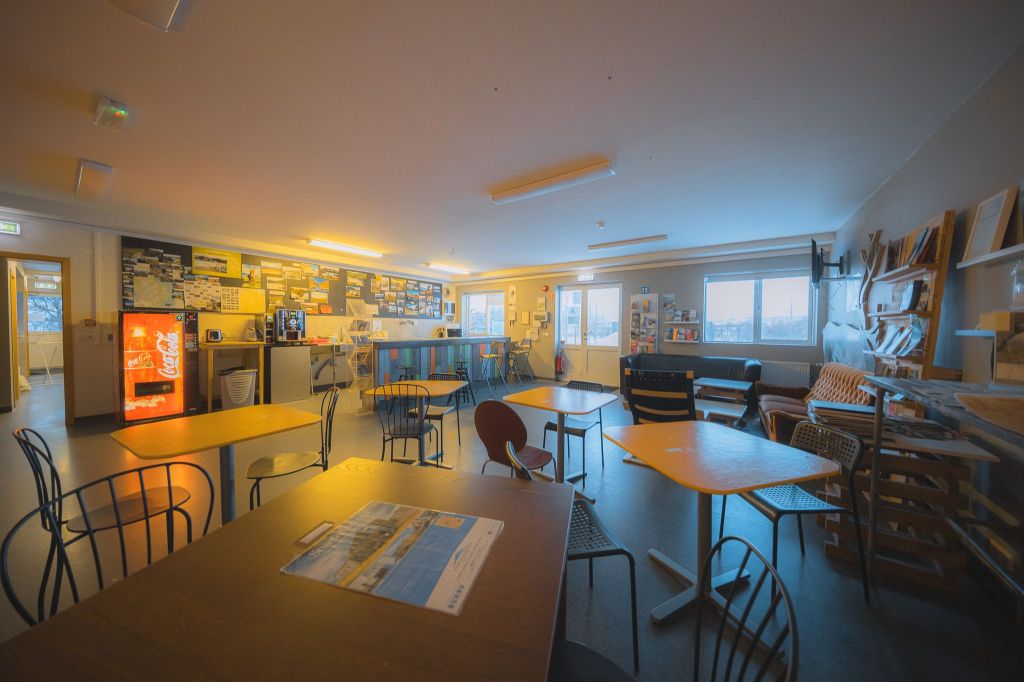 Cozy lounge with warm lighting, featuring mismatched tables and chairs, a vibrant red vending machine, and large noticeboards.