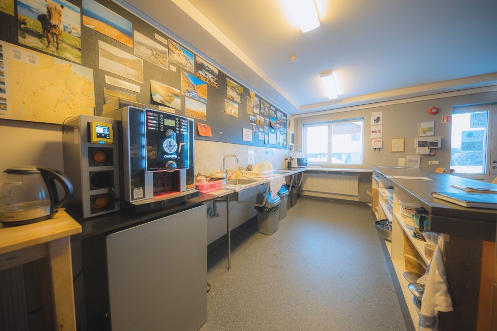 Kitchen area with a coffee machine, kettle, and sink on the left. Brightly lit room with large window, wall art, and shelves on the right.