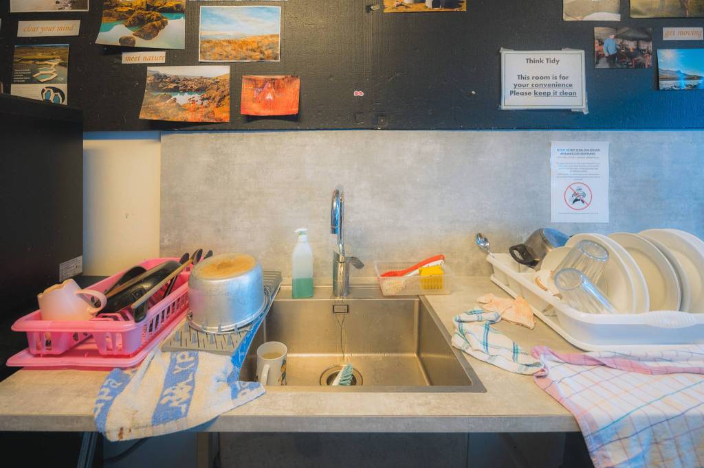A kitchen sink with a dish rack on each side. On the left, pink rack holds utensils and cups; on the right, a white rack with plates. Signs and photos adorn the wall above.