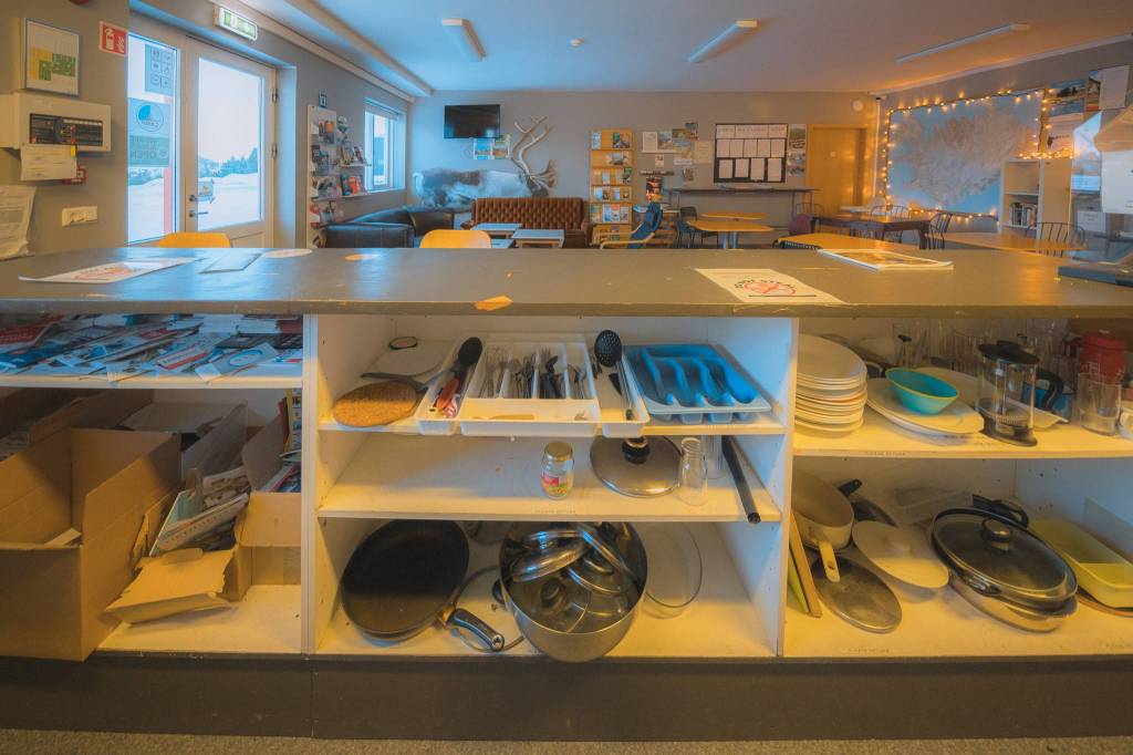Cozy room with a kitchen counter displaying utensils, plates, and pans. Warm lighting, seating area, and a map create a welcoming atmosphere.