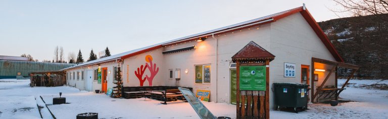 A long, single-story building with a red roof in a snowy landscape. A colorful mural of a hand and sun decorates the wall. Pine trees are visible in the background.