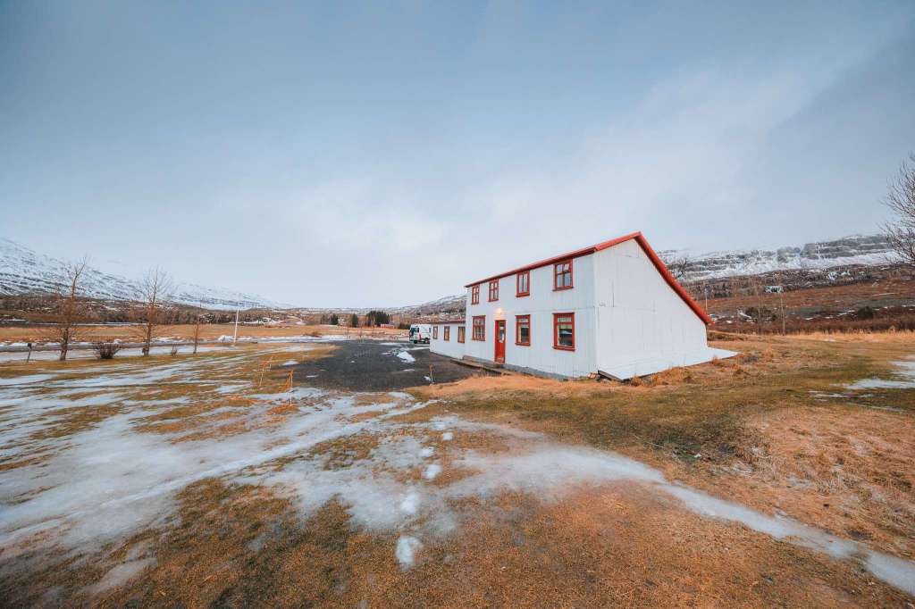 A white house with red trim is set against a snowy, mountainous landscape under a cloudy sky. Patches of snow dot the grassy terrain.