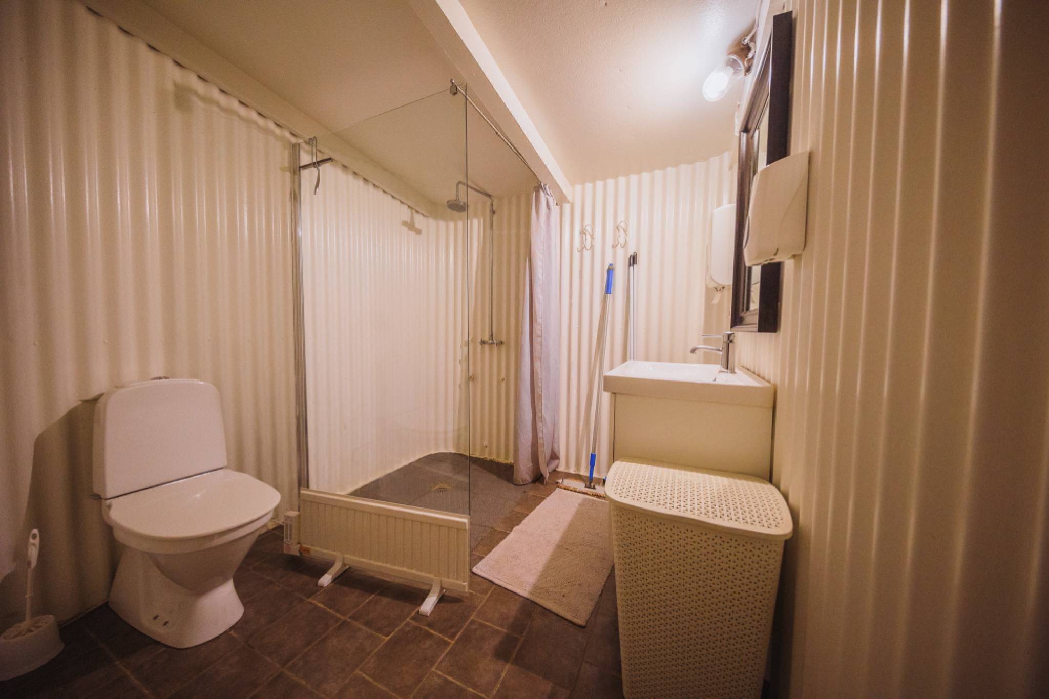 Minimalist bathroom with corrugated metal walls, featuring a glass shower, toilet, sink with mirror, laundry basket, and earthy-toned flooring.