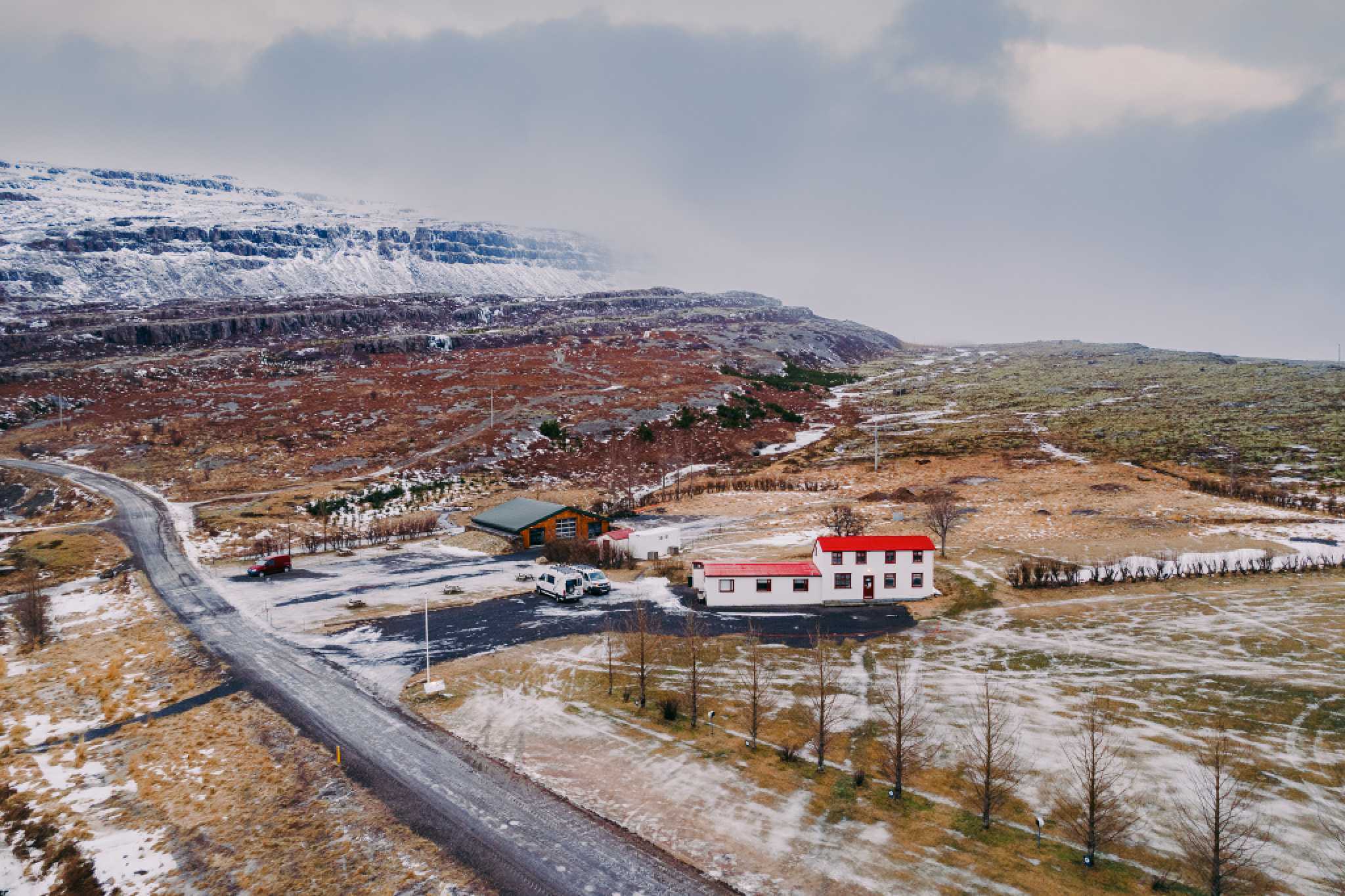 Aerial view of a snow-dusted landscape with a white house and red roof beside a road. Surrounding hills are lightly frosted under an overcast sky.