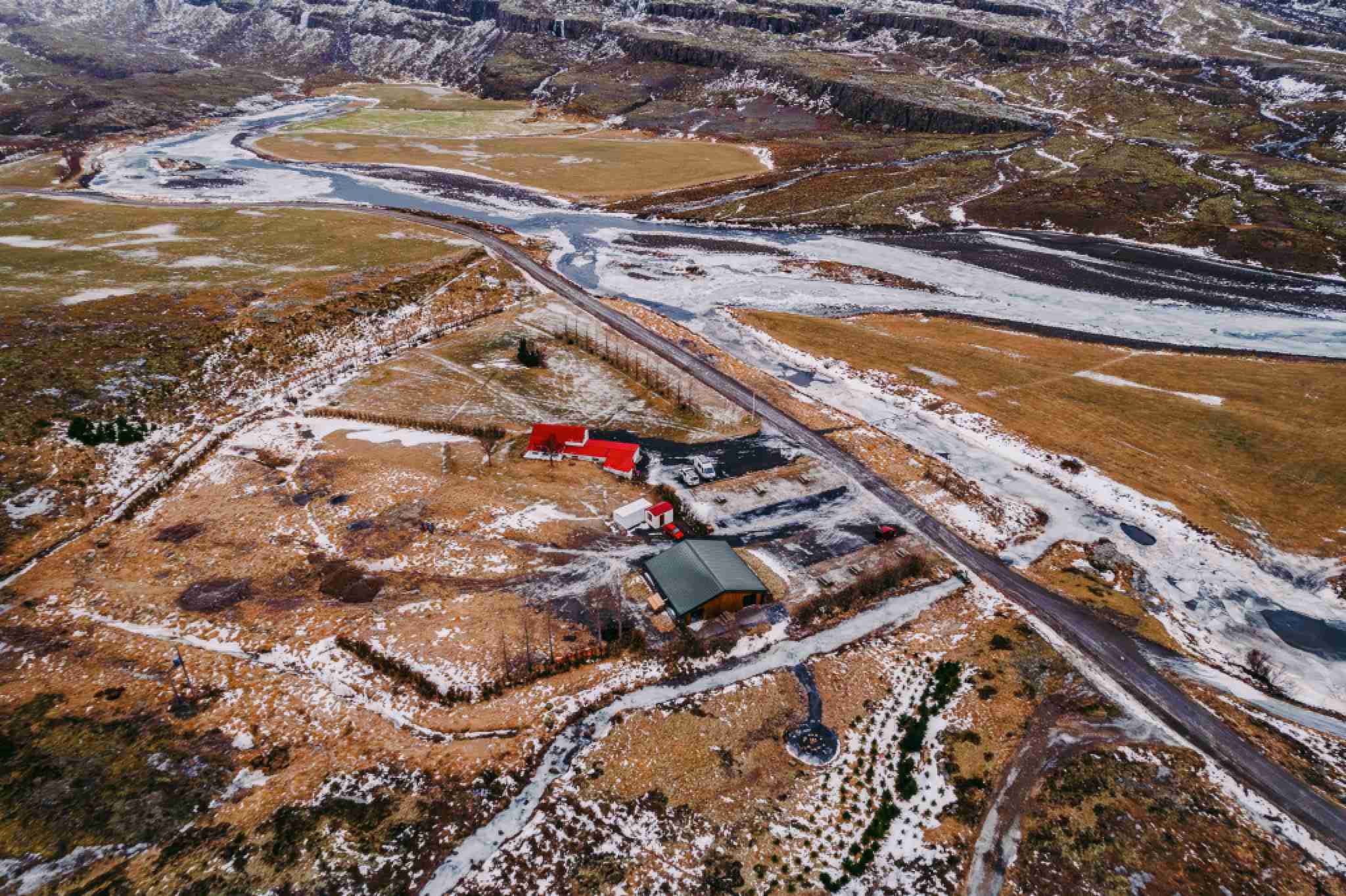 Aerial view of a snowy landscape with a winding river and brown fields. A road divides the scene, with red-roofed buildings nearby.
