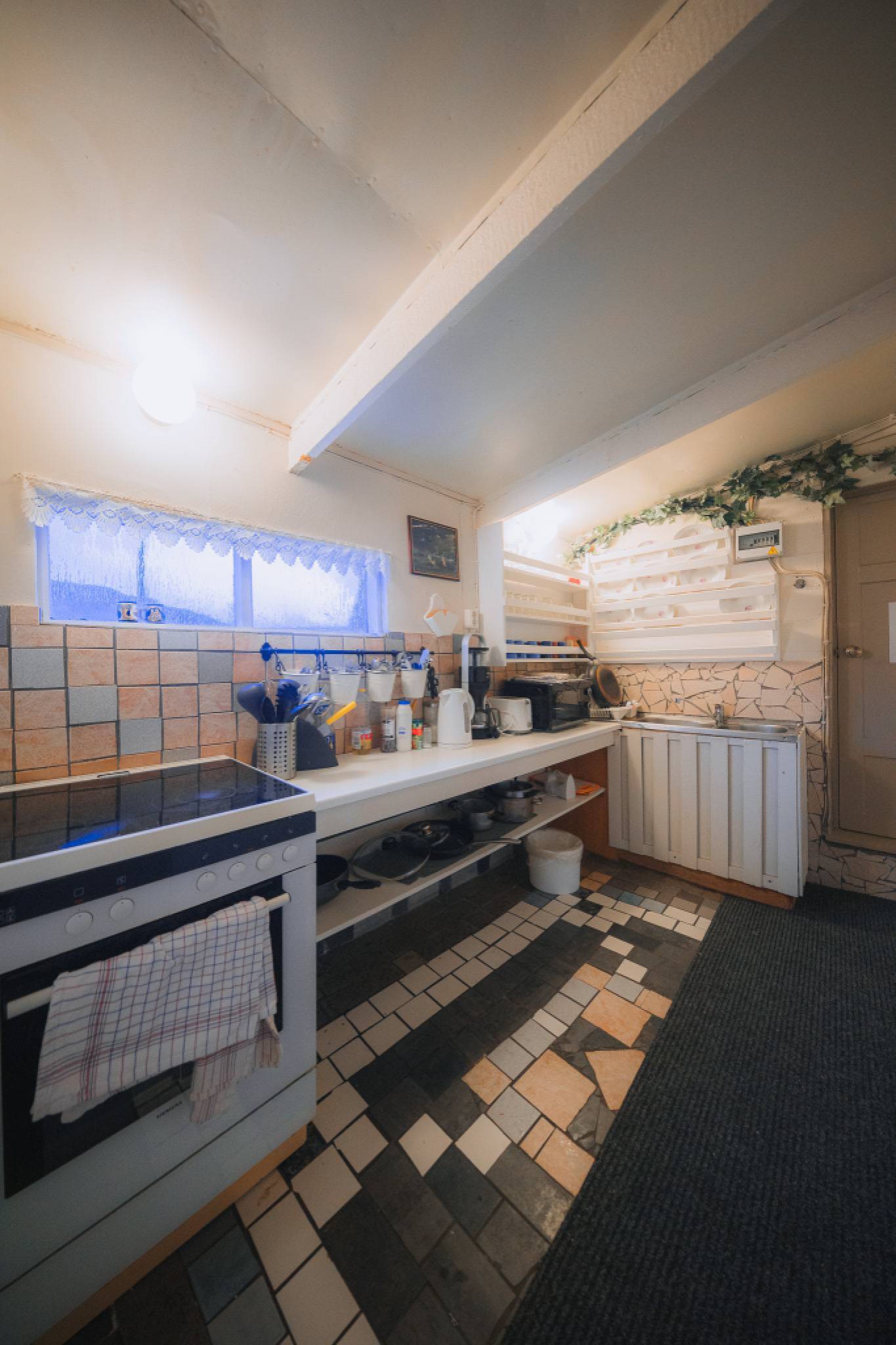 Cozy vintage kitchen with tiled floor, white countertops, and a rustic stove. A small window with lace curtains adds charm.
