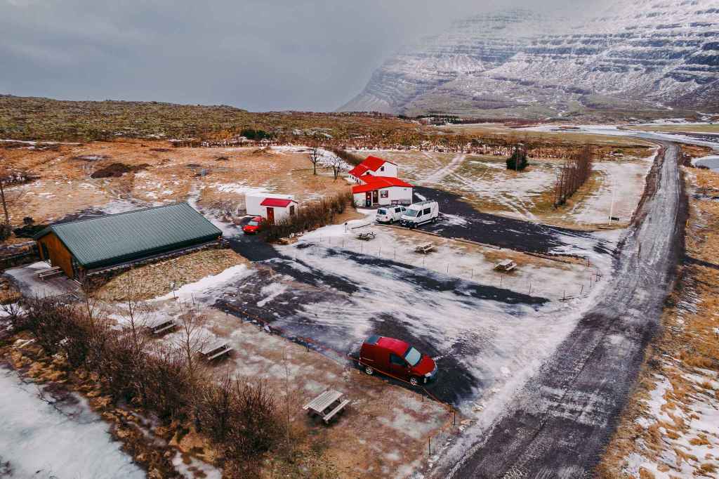 Aerial view of an icy parking lot with a red car, two white campers, and buildings with red roofs. Surrounding landscape of snowy mountains and grass.