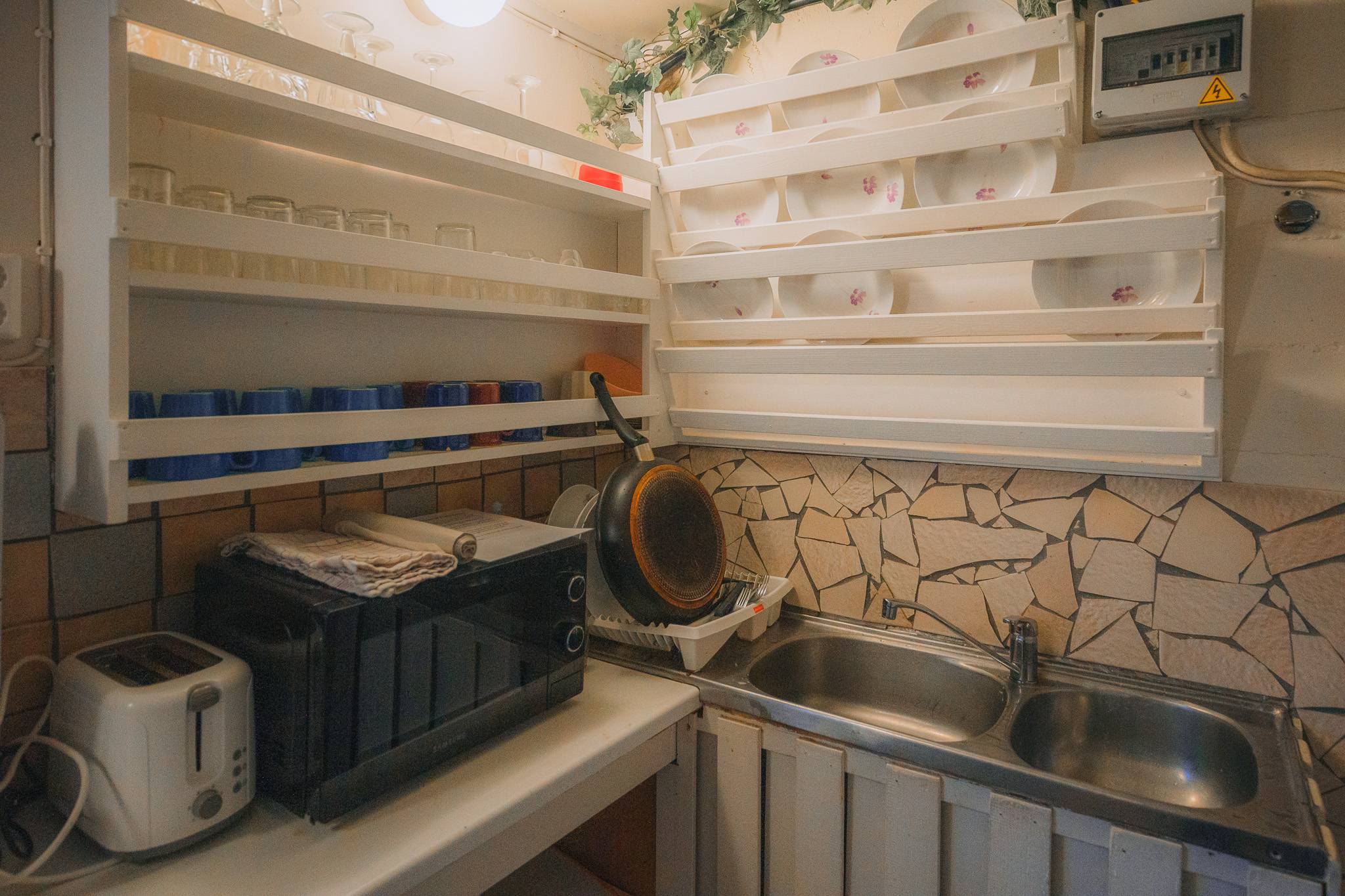 A cozy kitchen corner with white shelves holding glassware and plates. A toaster and microwave sit on the counter beside a double sink.