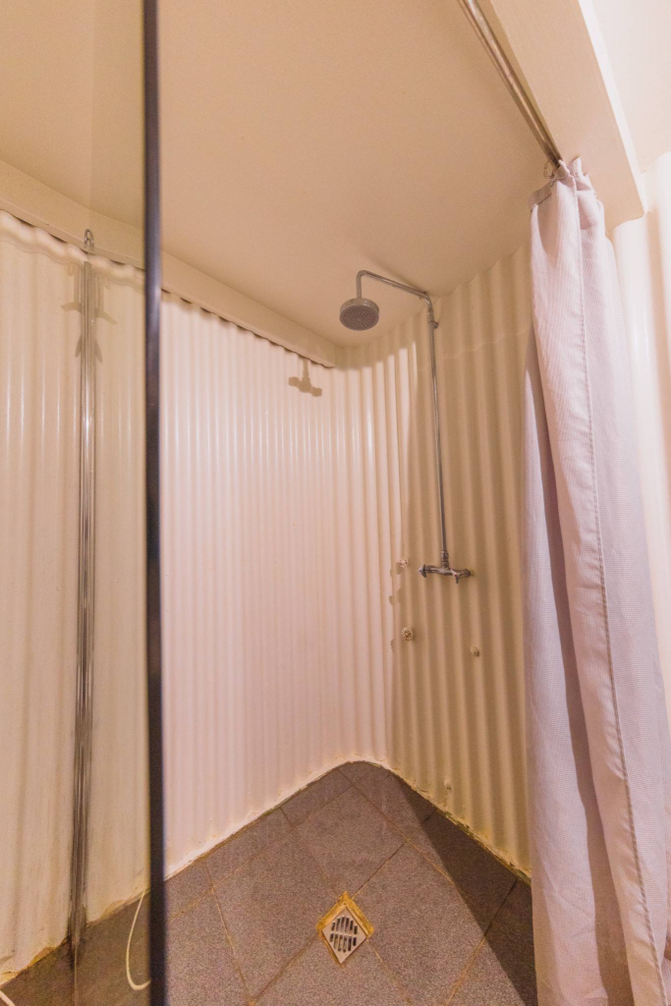 Minimalist shower with textured beige walls, a circular showerhead, and a white curtain on the right. Square floor drain visible.
