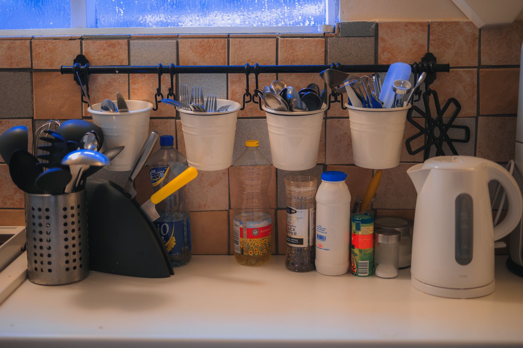 A cozy kitchen counter with hanging white containers holding cutlery, utensils in a holder, a white kettle, and various condiments against tiled walls.