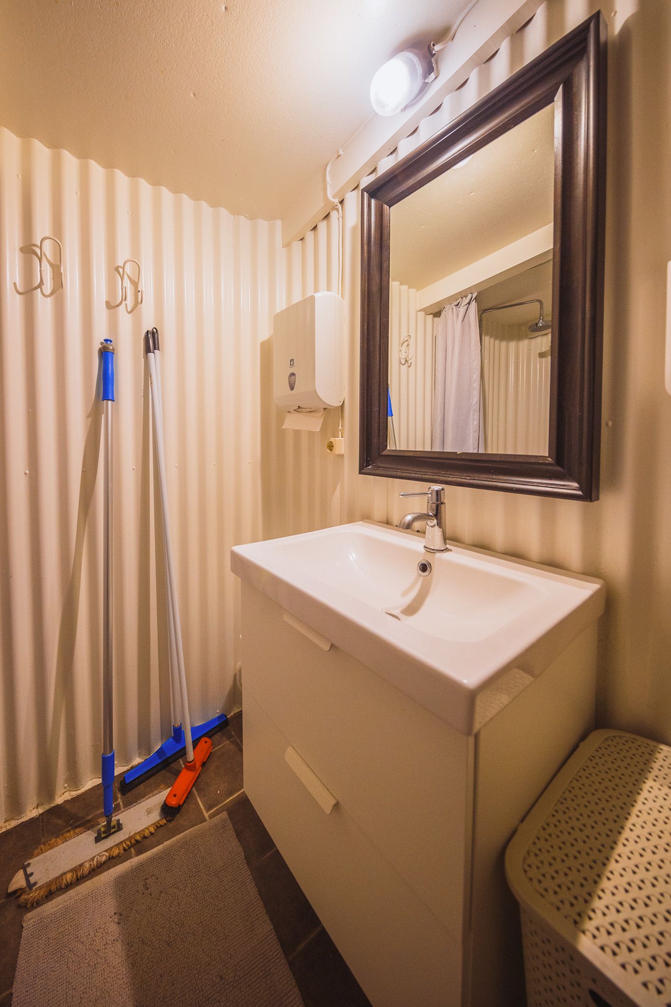 Small, well-lit bathroom with a white sink and dark-framed mirror. Cleaning tools lean against corrugated walls.
