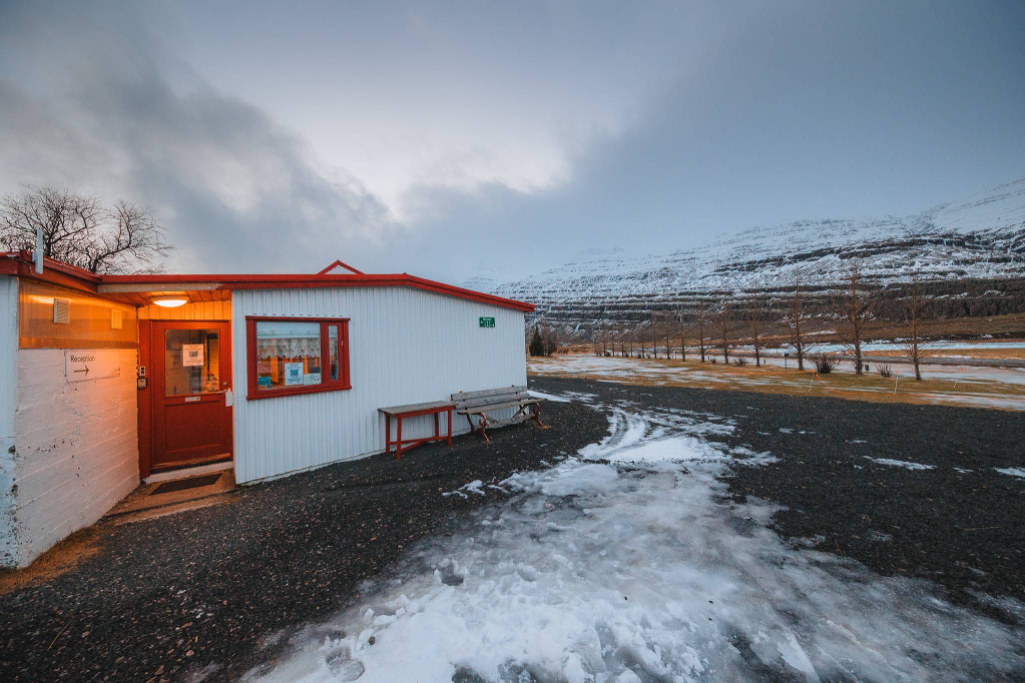 Small white cabin with a red roof stands in a snowy landscape. Warm light glows from the window. Snow-covered mountains in the background under a cloudy sky.