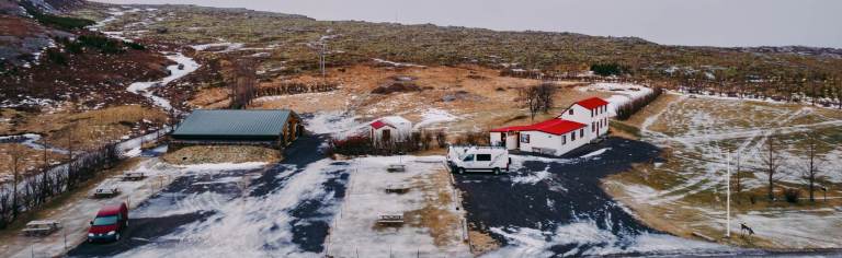 Aerial view of a snowy landscape with two white buildings featuring red roofs. A white van and red car are parked nearby. Brown grassland surrounds the scene.