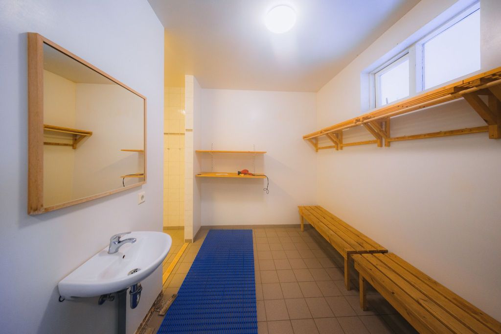 Clean locker room with a sink, mirror, and wooden benches. Shelves on the wall, bright lighting from a window, and a blue mat on tiled floor.