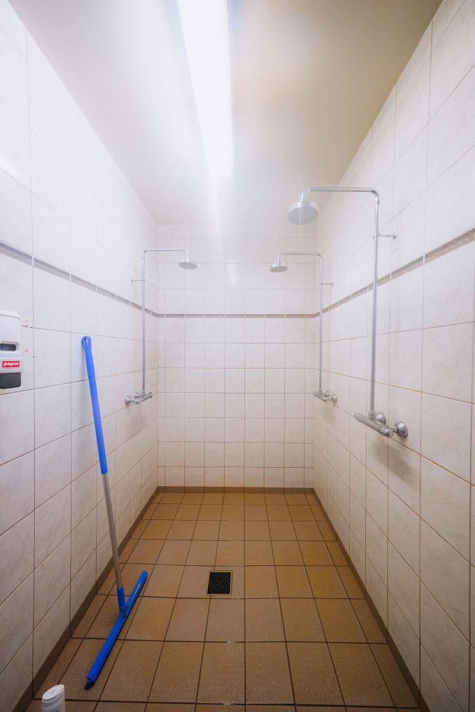 Public shower stall with white tiled walls and brown tiled floor. Three showerheads are visible, with a blue mop and a soap dispenser on the left.