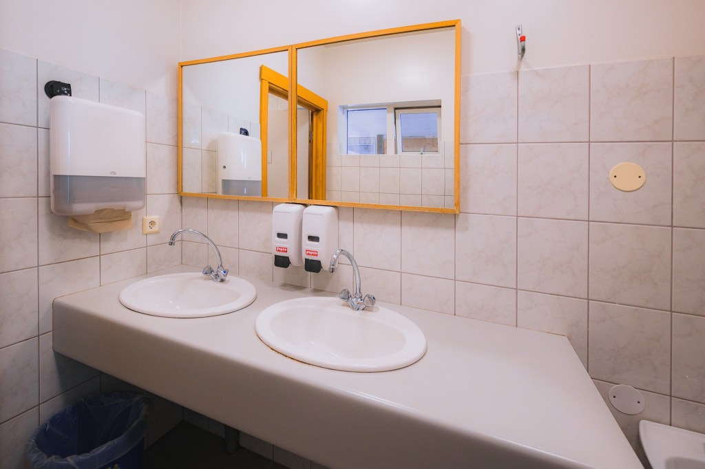 A clean, tiled restroom features a double sink with two faucets, soap dispensers, and a large mirror with a bright yellow frame.