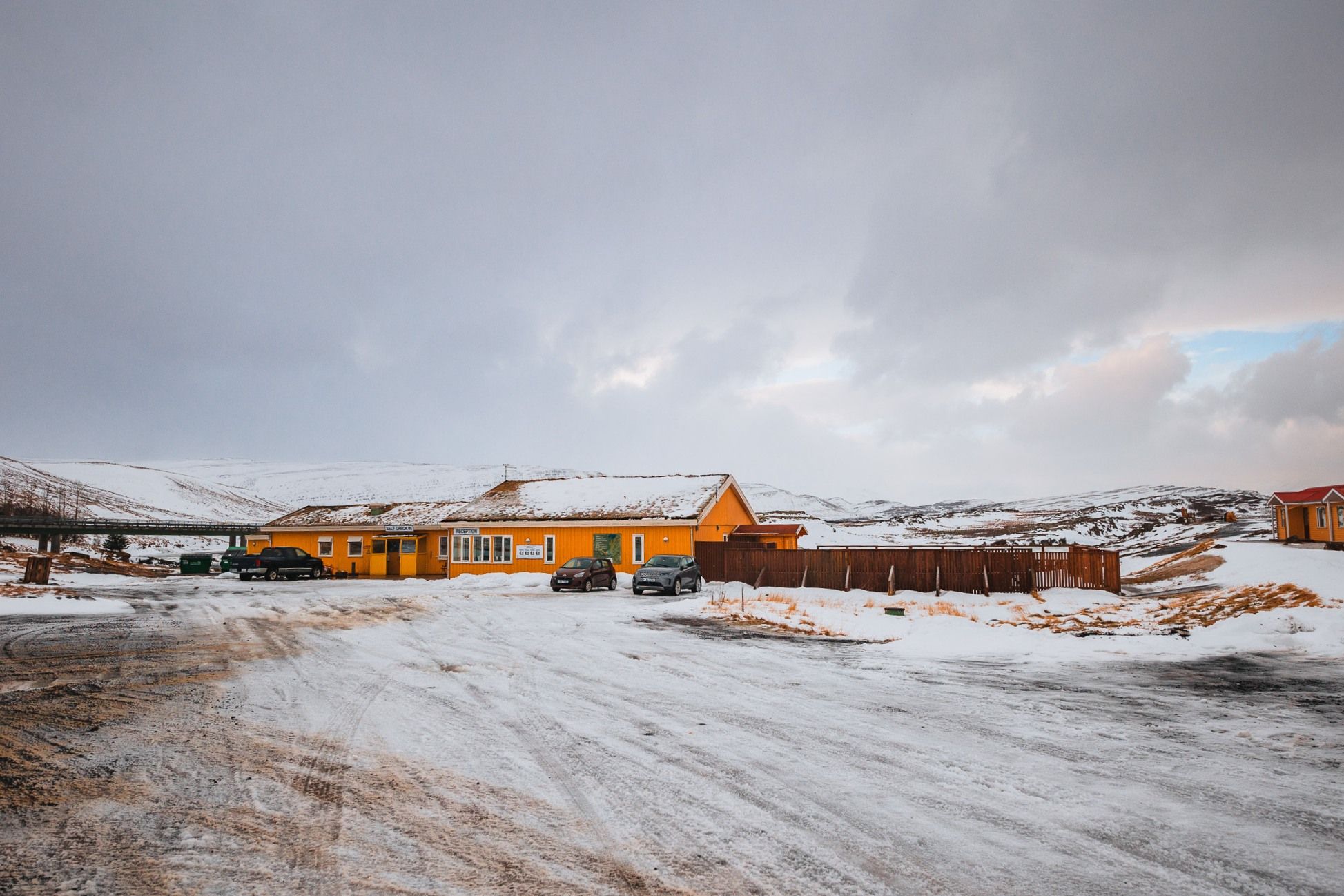 Yellow building set in a snowy landscape under a cloudy sky. Several cars are parked nearby.