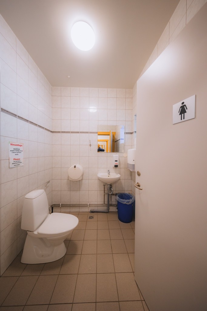 A clean, well-lit public restroom with a toilet, sink, and mirror. The beige tiles create a sterile feel. A door sign indicates it's a women's restroom.