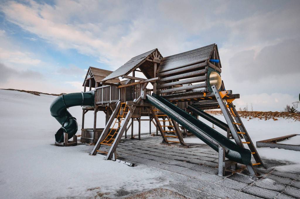 Snow-covered playground equipment with slides and ladders stands in an empty field under a cloudy sky.