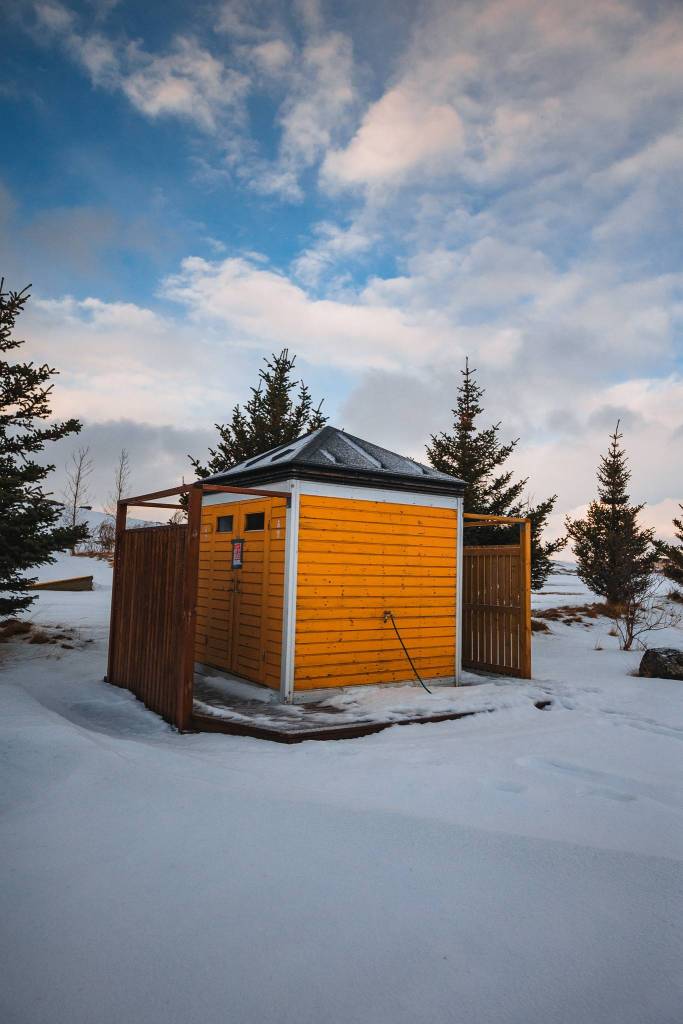 A small yellow wooden cabin stands in a snowy landscape, surrounded by evergreens under a partly cloudy blue sky.