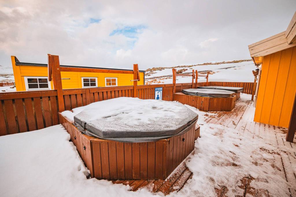 Snow-covered hot tubs on a wooden deck, surrounded by a yellow cabin and snowy landscape.