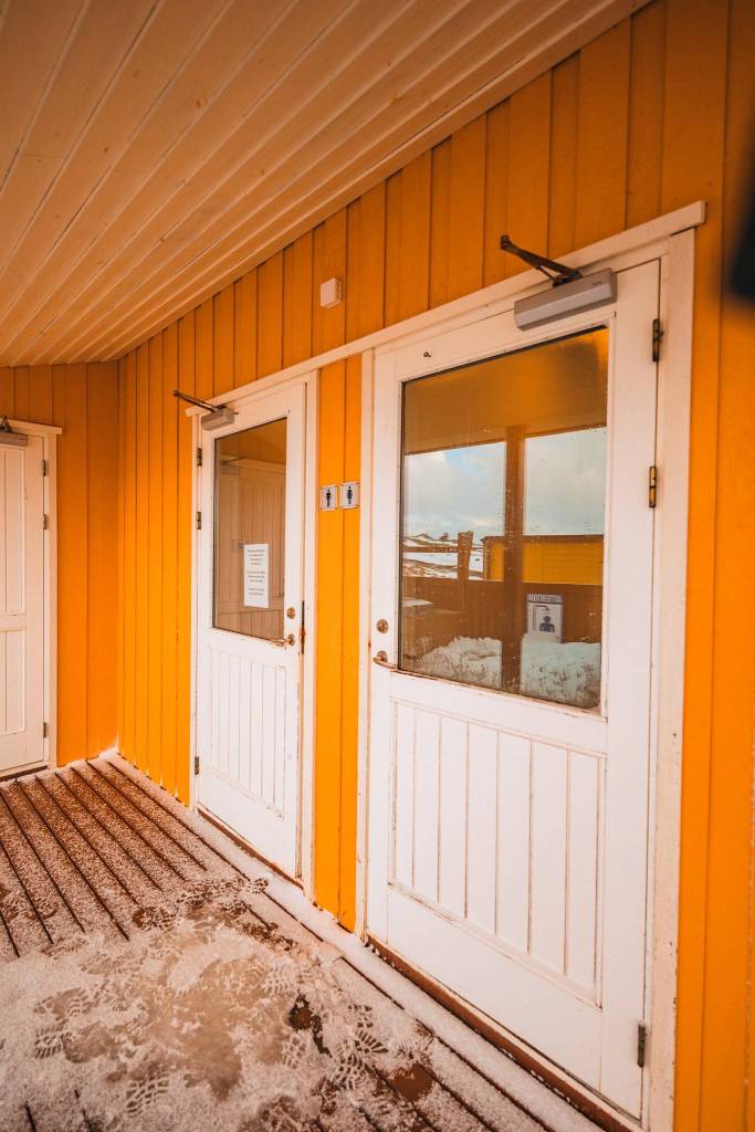 Yellow wooden building exterior with two white doors, snowy footprints on the wooden floor, and a reflected snowy landscape in the door windows.