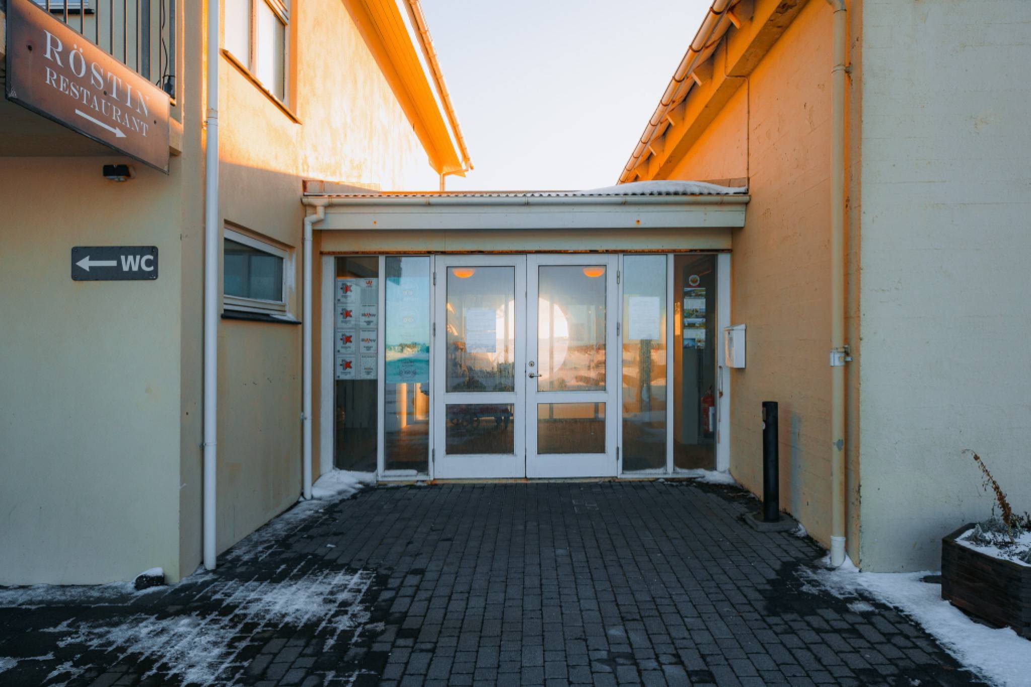 Entrance of a light-orange building with glass double doors, leading to a restaurant. A sign points left to the restroom. Some snow is on the ground.