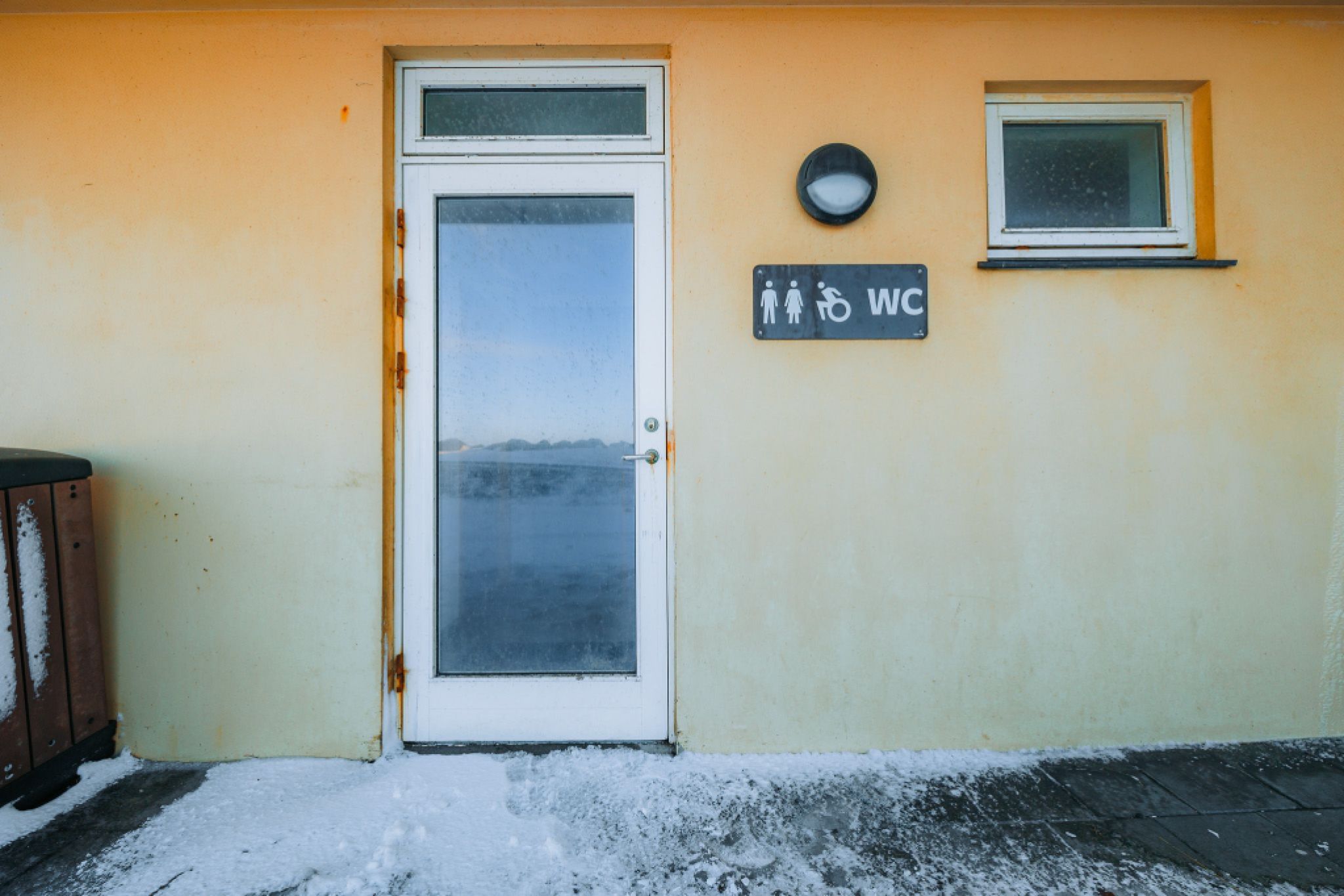 Frost-covered public restroom entrance with a glass door, located on an icy path. Signage indicates accessibility and unisex use.