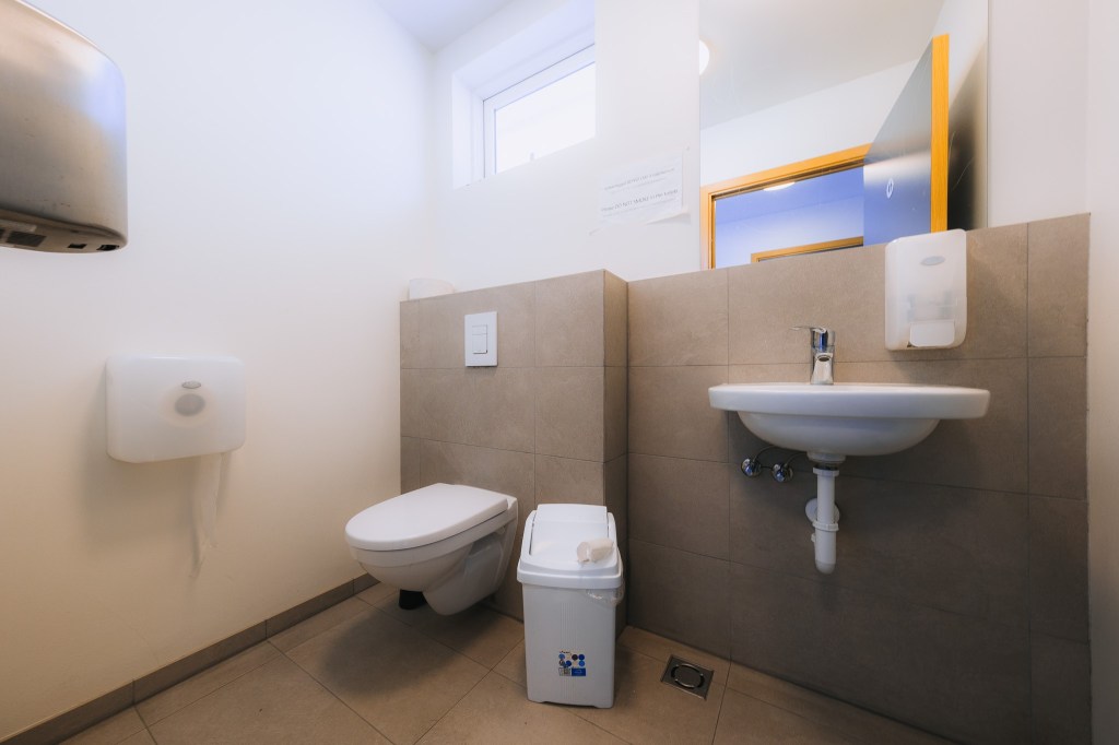 Clean public restroom with tiled walls, featuring a wall-mounted toilet, sink with a soap dispenser, small bin nearby, and bright natural lighting.