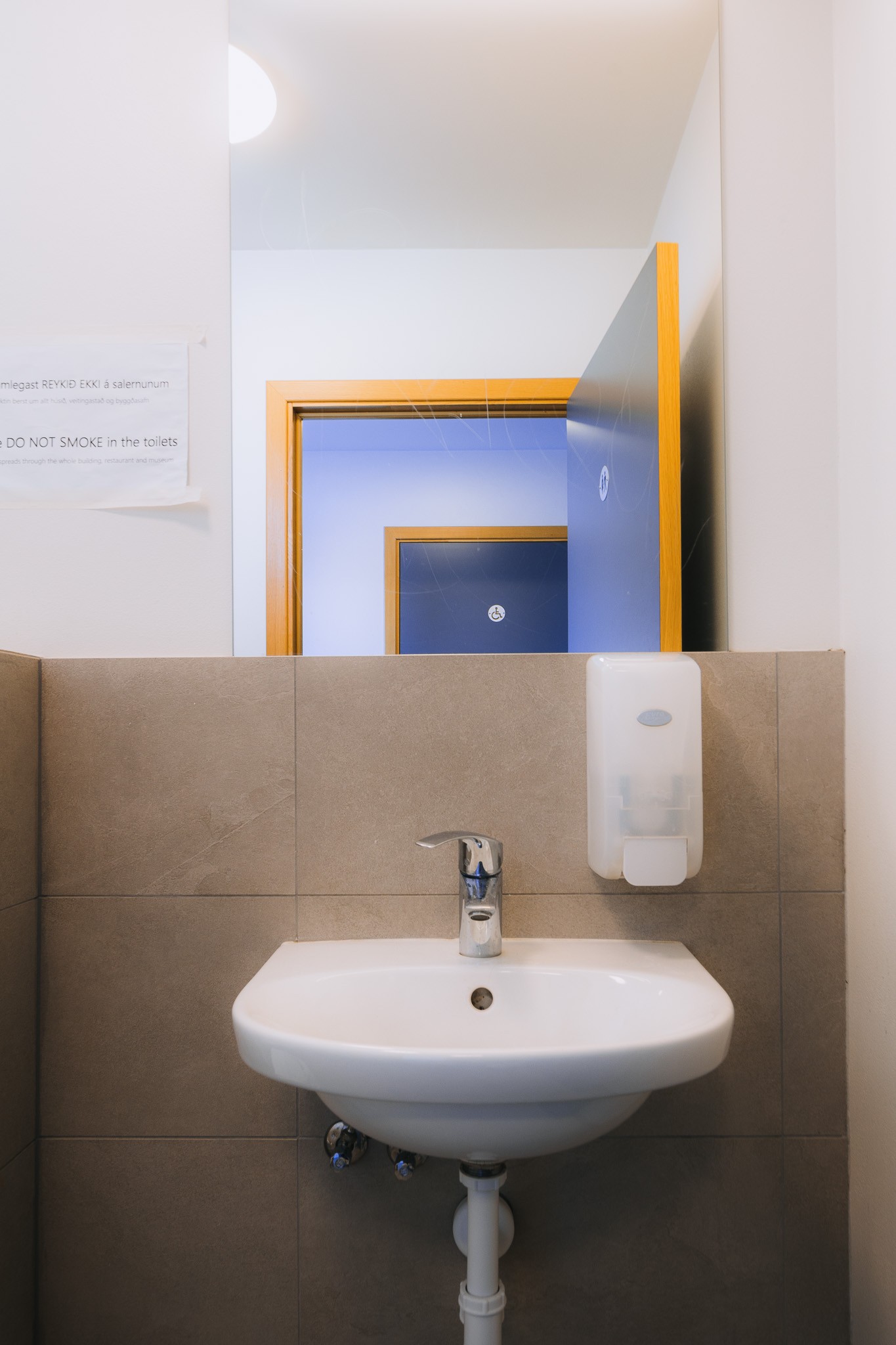 A small restroom with a white sink, silver faucet, and wall-mounted soap dispenser. A mirror reflects blue doors.