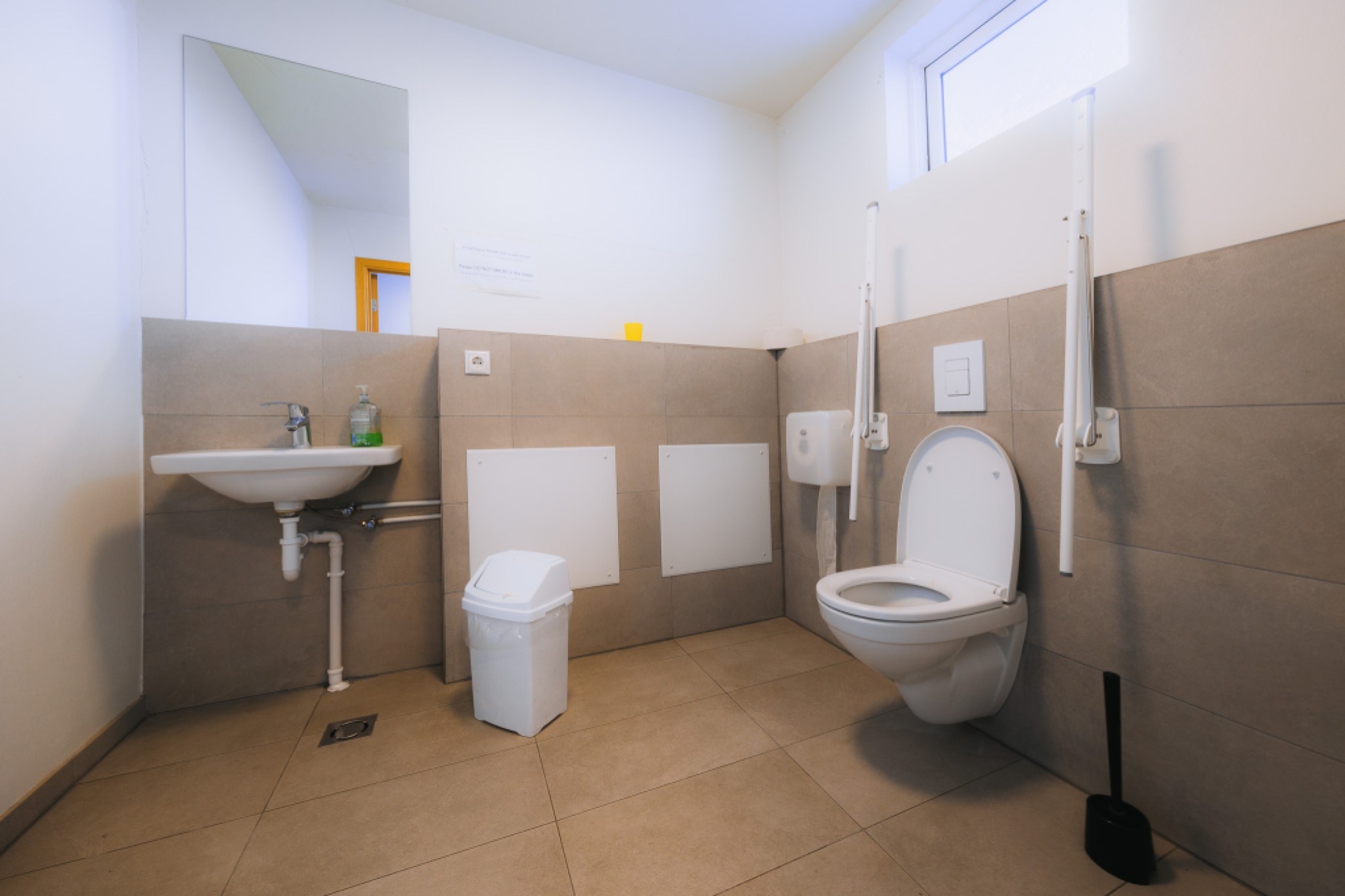 Accessible bathroom with a wall-mounted toilet, support rails, a small sink with soap, a white trash bin, gray tiles, and a window for natural light.