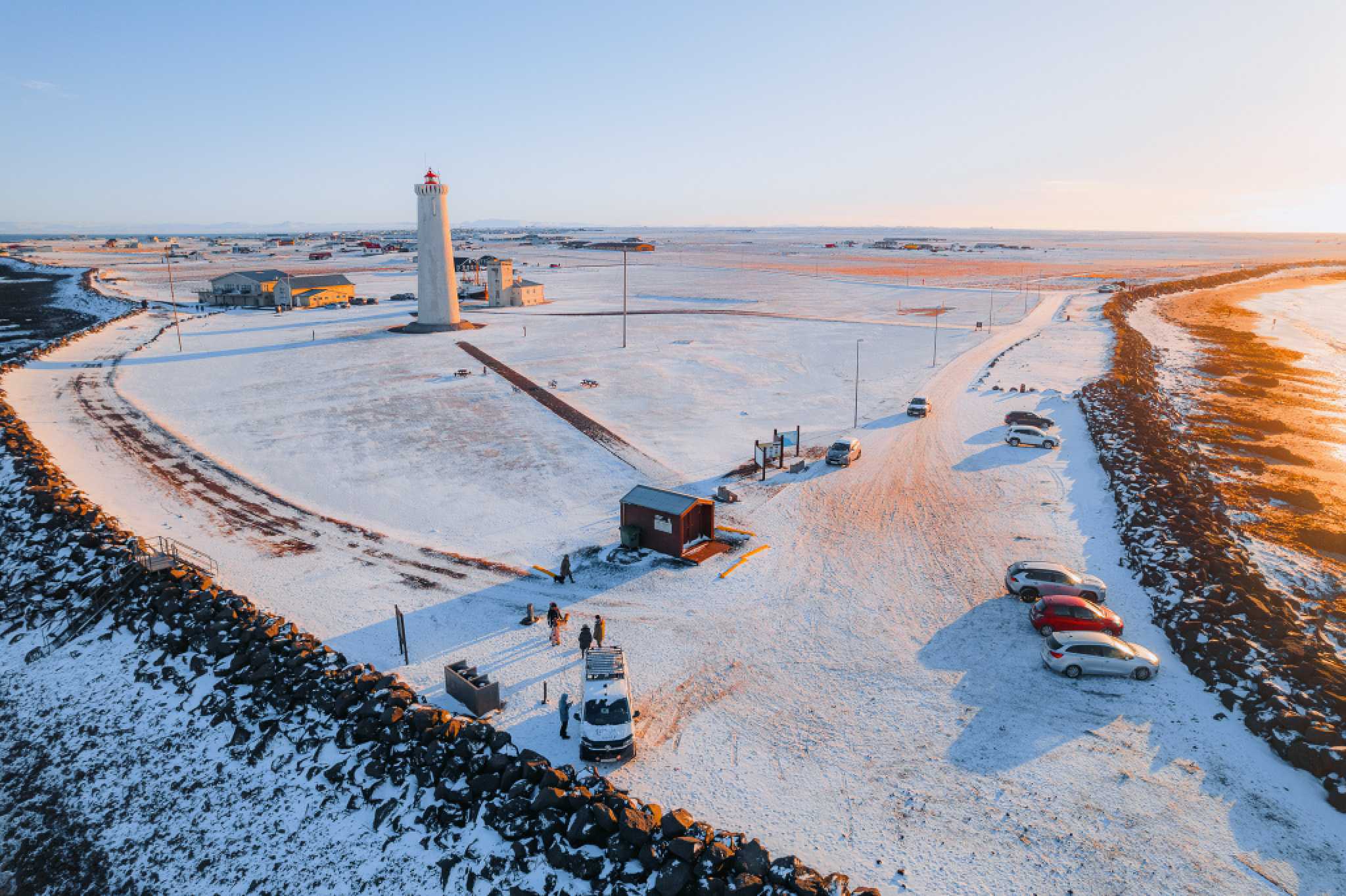 A snowy landscape with a tall, white lighthouse in the center. Cars are parked nearby, and a few people walk on the icy ground.