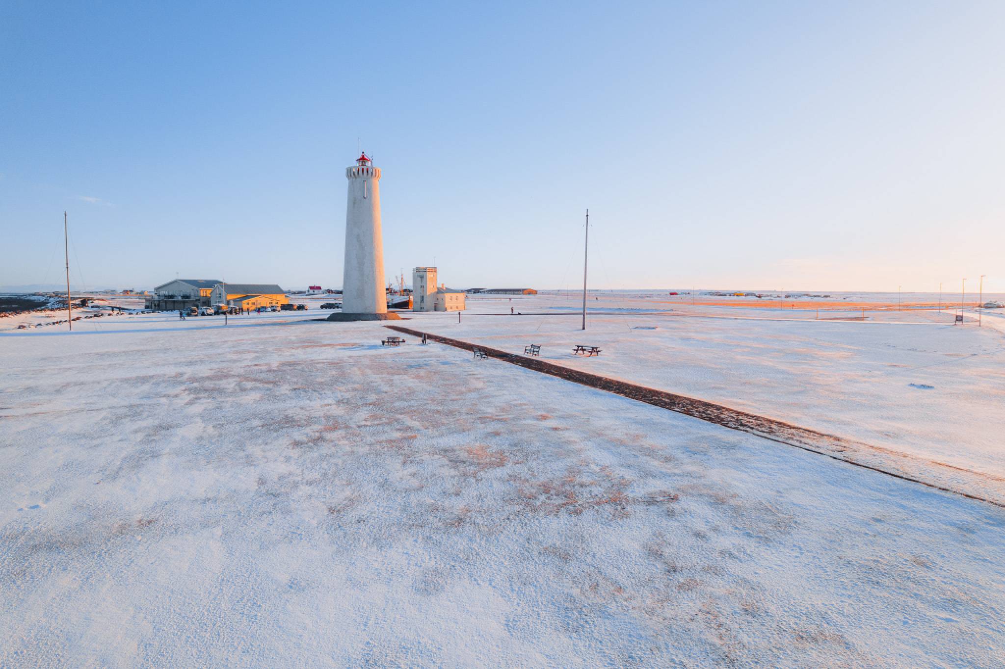 Tall lighthouse on a snowy landscape at sunrise, casting long shadows. Nearby are small buildings and benches.