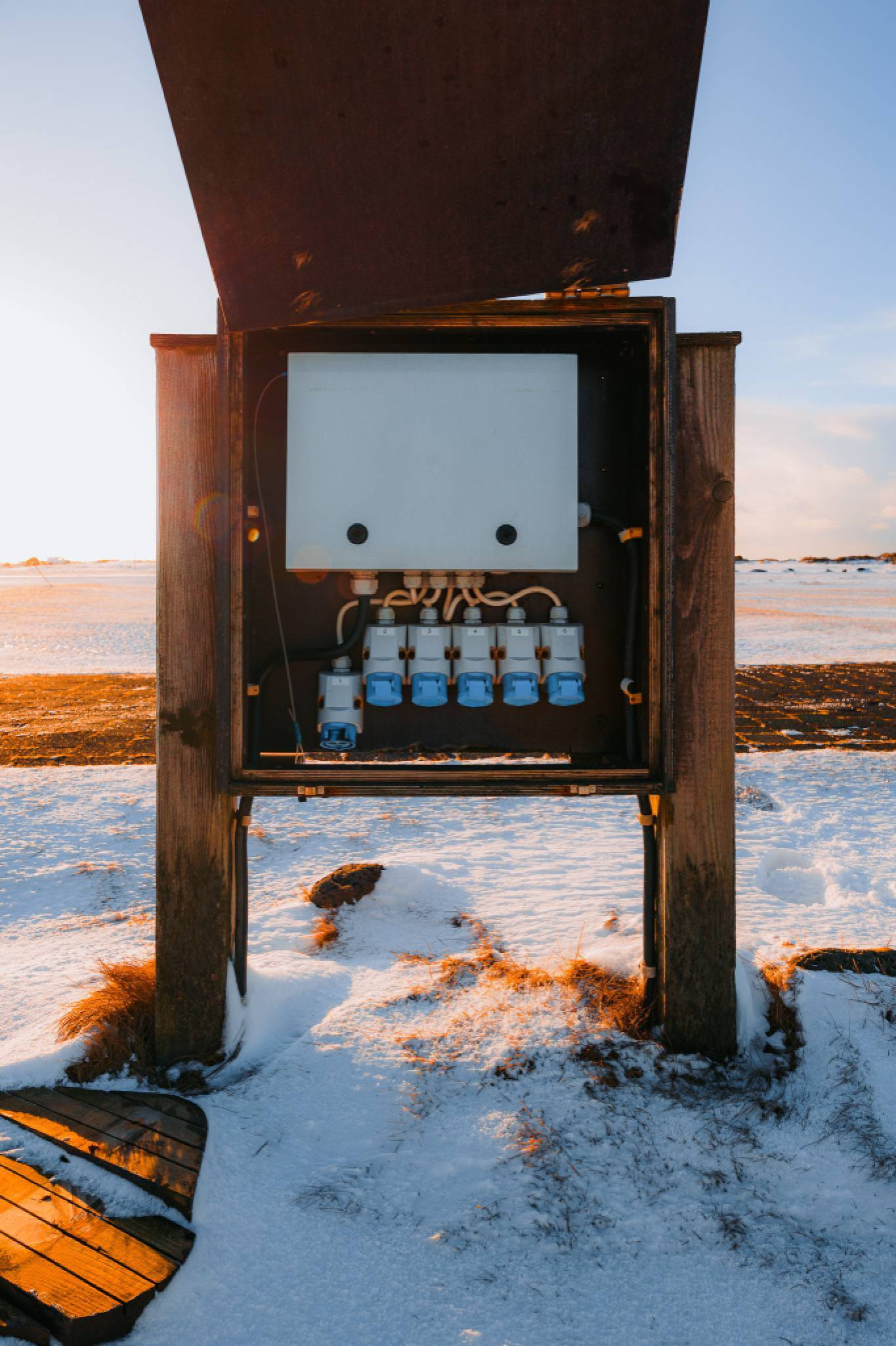 Wooden power box with open cover in snowy landscape at sunset. Blue electrical sockets and tangled cables inside.