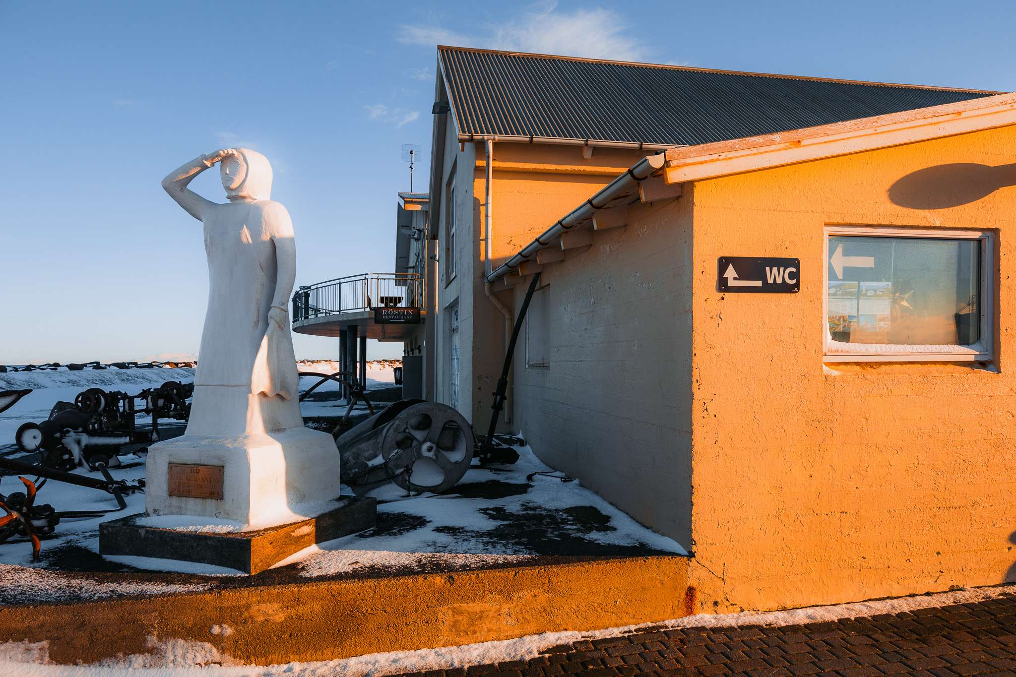 A white statue of a person shielding their eyes stands next to a yellow building with a "WC" sign. Snow covers the ground, under a clear blue sky.
