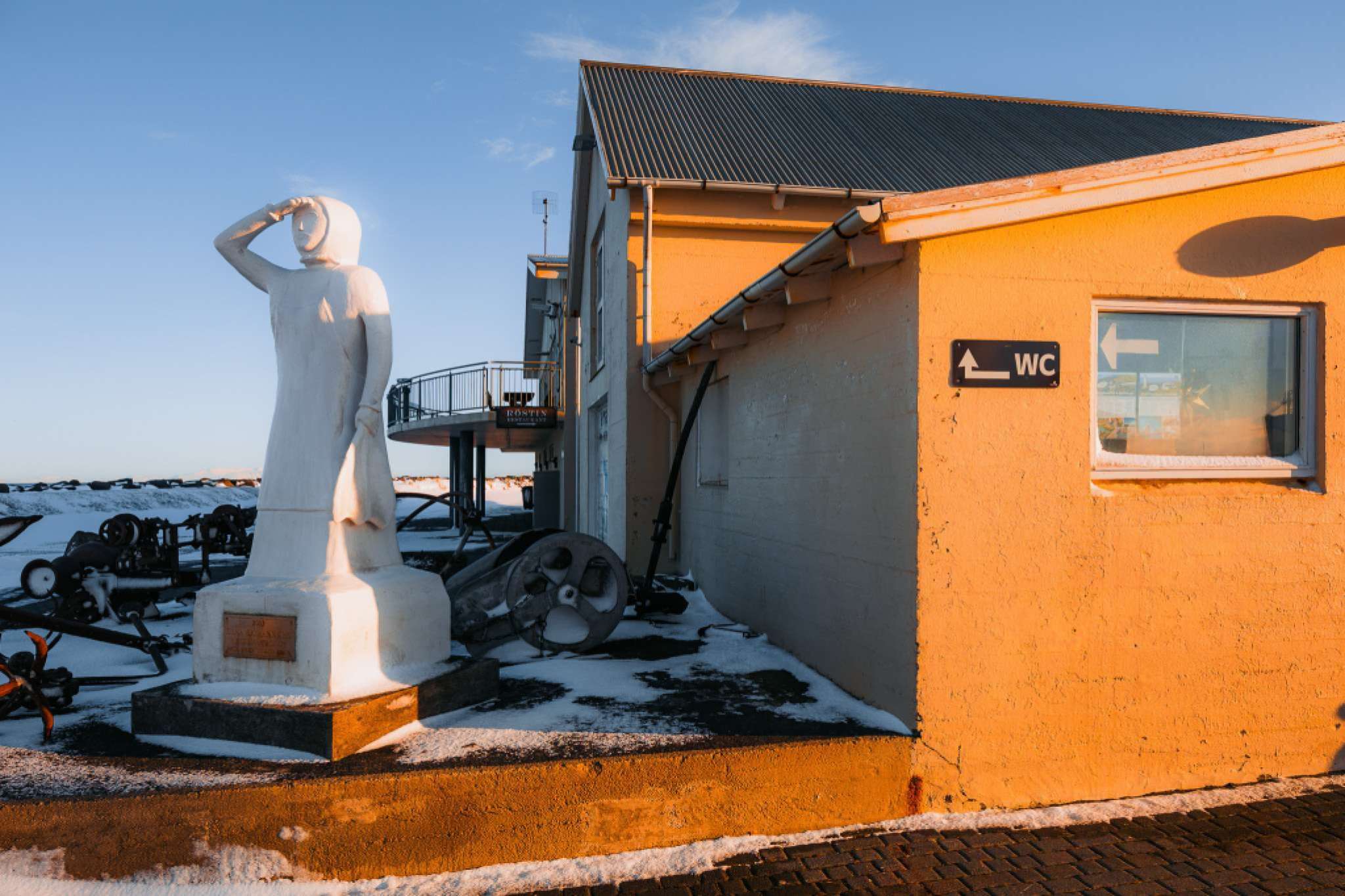 A white statue of a person shielding their eyes stands next to a yellow building with a "WC" sign. Snow covers the ground, under a clear blue sky.
