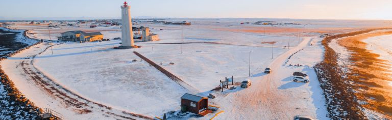 Aerial view of a snowy landscape with a tall lighthouse at the center, surrounded by small buildings and scattered cars.