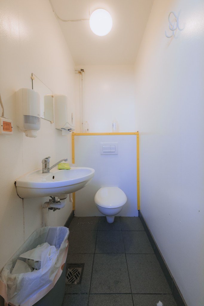 A small, brightly lit bathroom with white walls features a wall-mounted sink and toilet. A trash bin is visible on the left, and dispensers are above the sink.