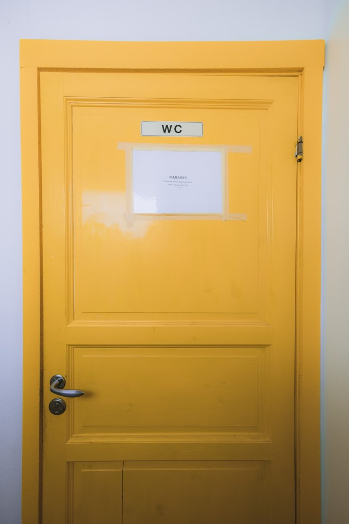 Yellow wooden door with a "WC" sign and taped white paper. The door has a silver handle and is set against a white wall.