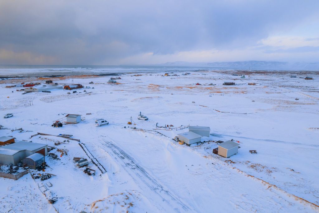 A snowy, rural landscape with scattered small buildings under a cloudy sky.
