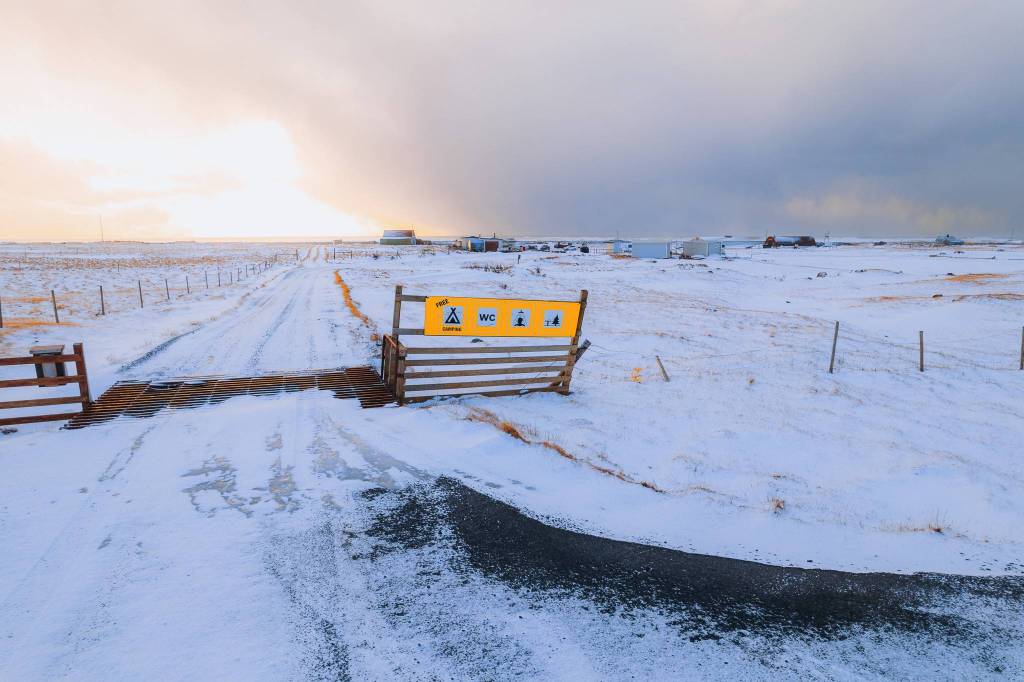 Snowy landscape with a dirt road leading to a horizon lit by a bright sky. A yellow sign shows camping and restroom symbols.
