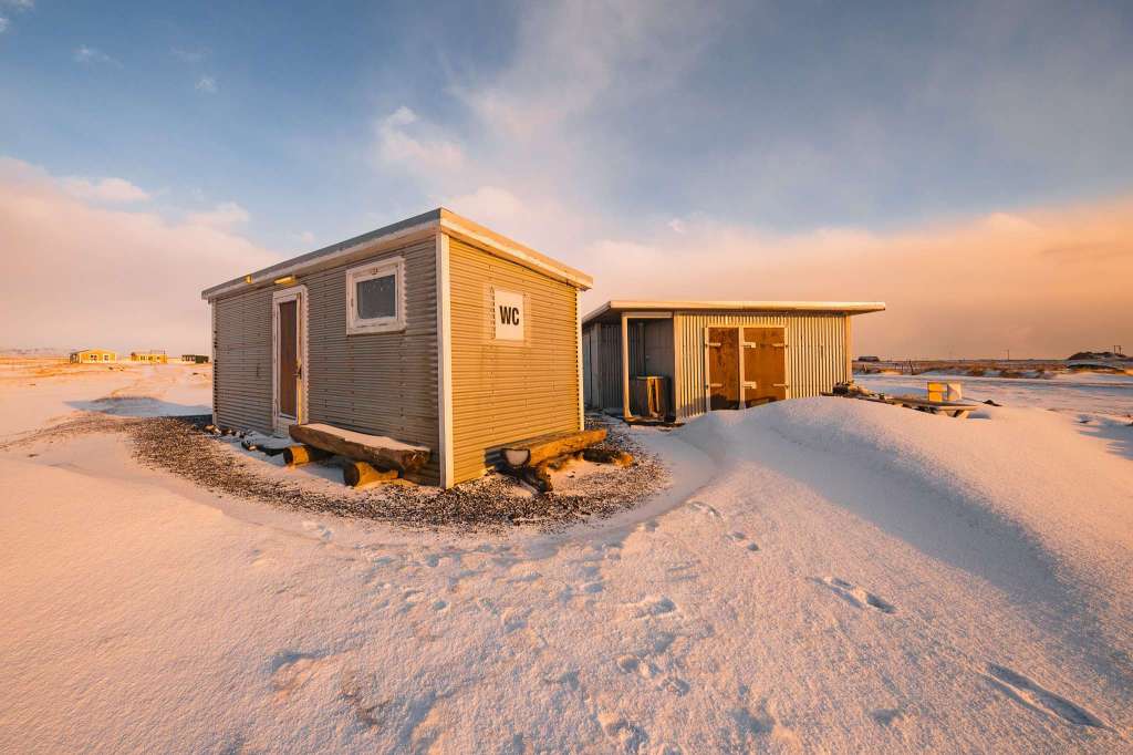 Two small, weathered buildings in a snowy landscape, one marked "WC." Golden sunlight casts warm tones.