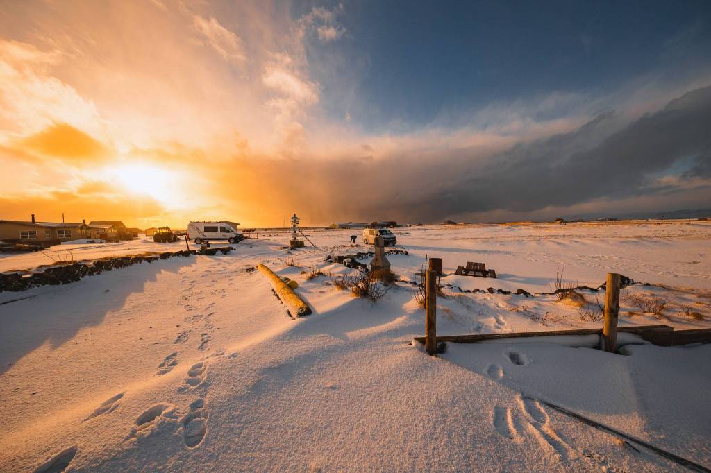 A snowy landscape at sunset, with footprints in the foreground leading to a parked camper van. The sky is vibrant with warm orange and yellow hues.