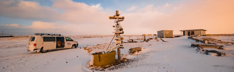 A snowy landscape features a white van near a wooden signpost with multiple directional arrows, small buildings in the background, and a dramatic sunset sky.