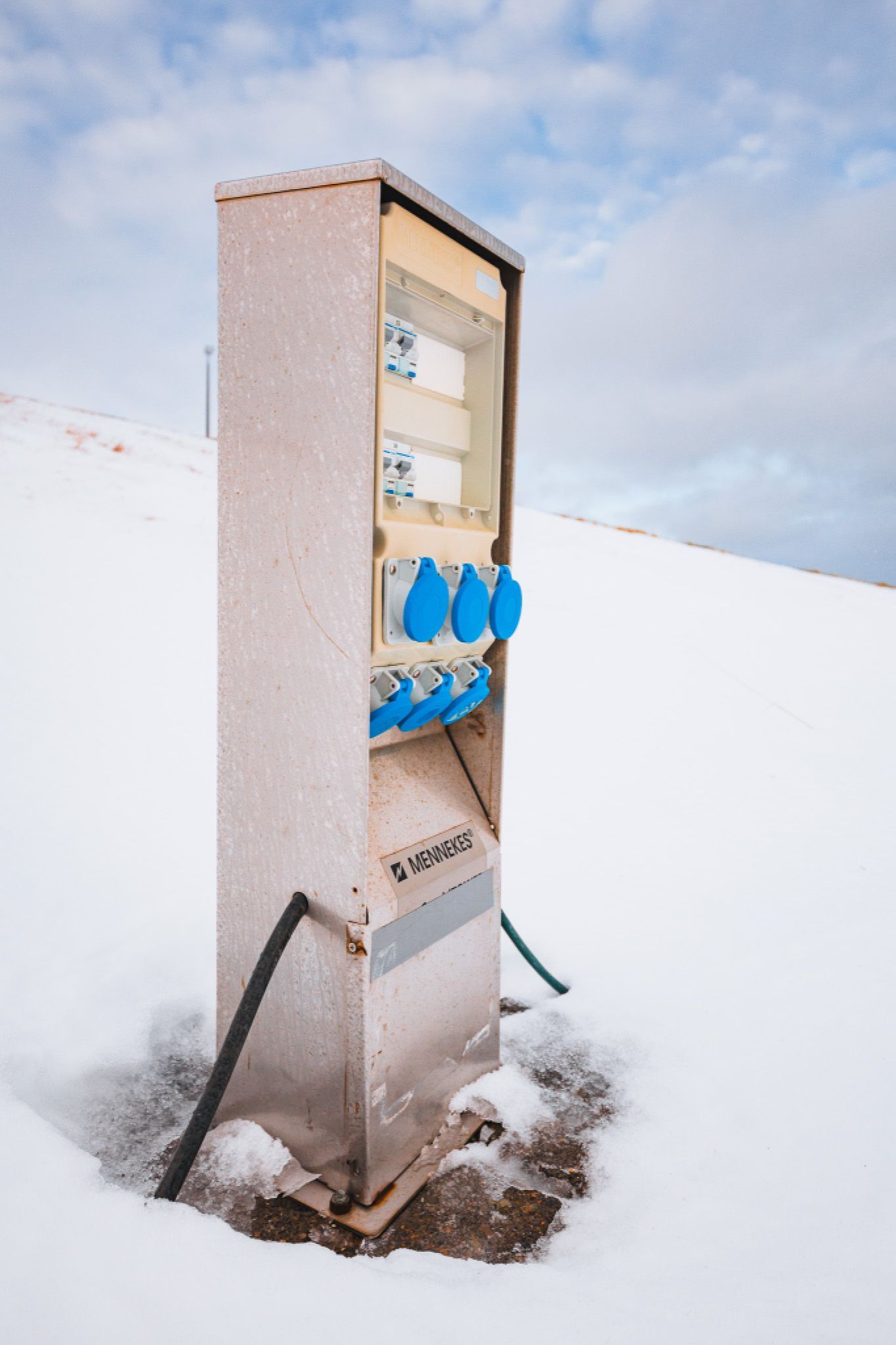 A metal electrical outlet stand with multiple blue sockets and black cords is set in a snowy landscape under a cloudy sky.