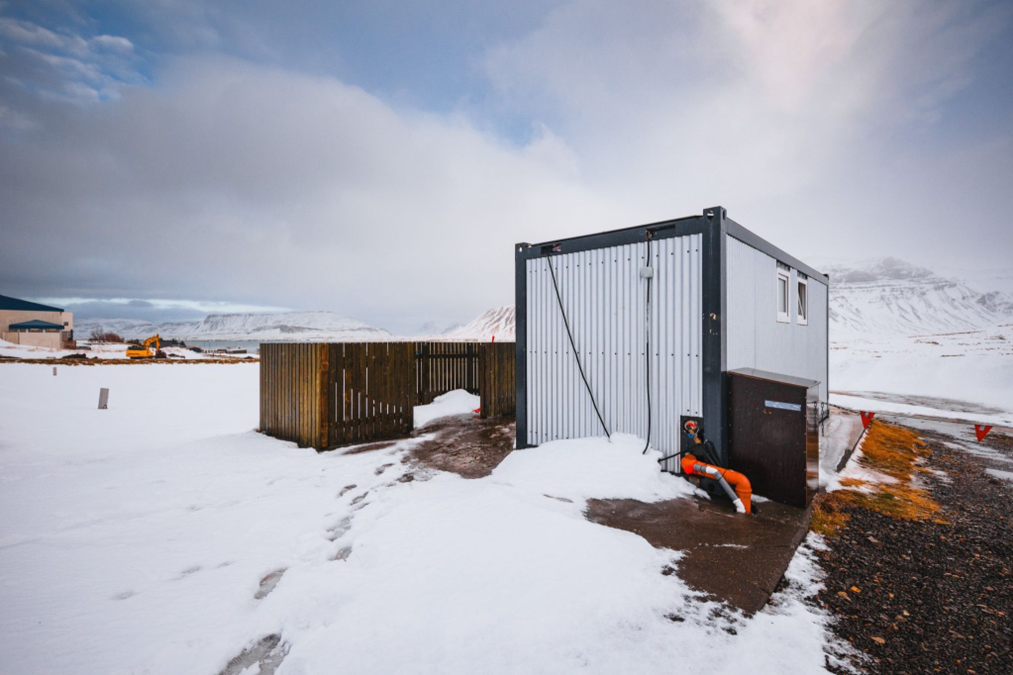A small, gray industrial building stands amid a snowy landscape with mountains in the distance under a cloudy sky.