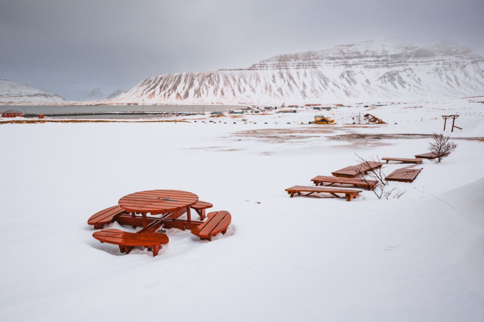 Snow-covered landscape with wooden picnic tables, set against a backdrop of icy mountains under a cloudy, gray sky.