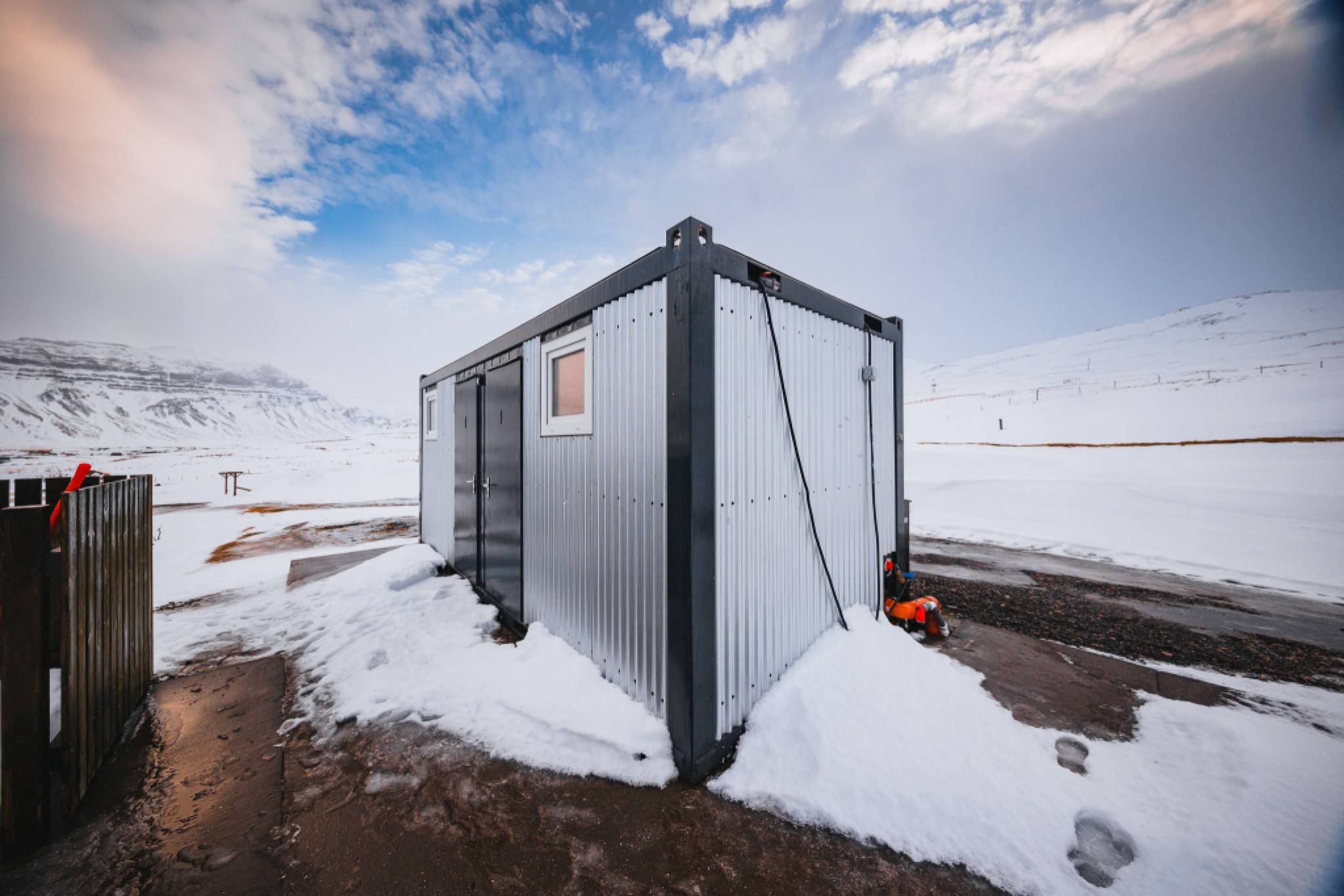 A small, metal cabin sits in a snowy landscape under a cloudy sky.