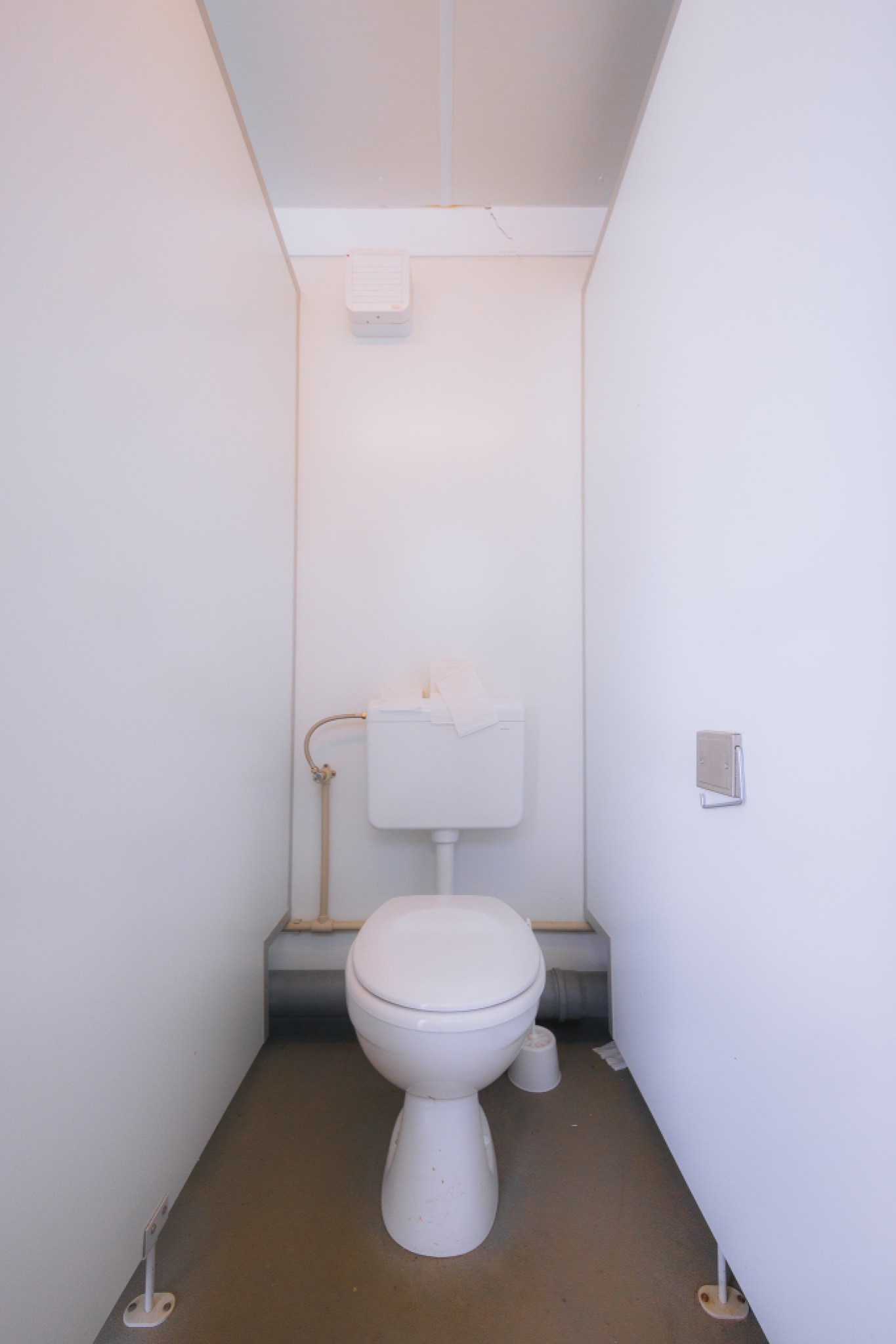 A small, minimalistic bathroom with white walls features a basic white toilet and tank. A piece of toilet paper rests on the tank.