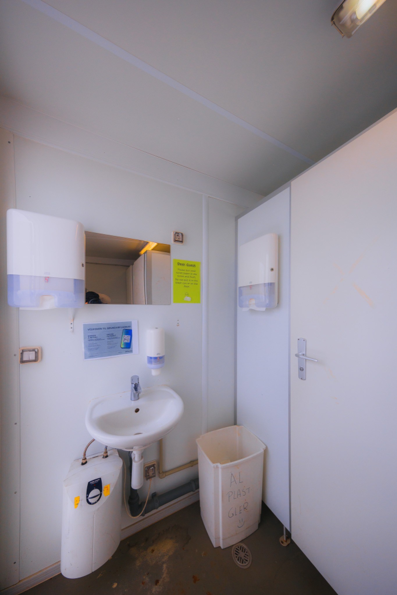A small, clean bathroom with white walls features a sink below a mirror, soap dispensers, a waste bin, and a closed door.